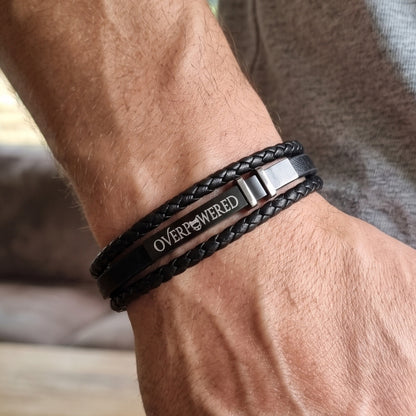 Black leather braided bracelet on wrist with silver-tone clasp and OVERPOWERED logo plate