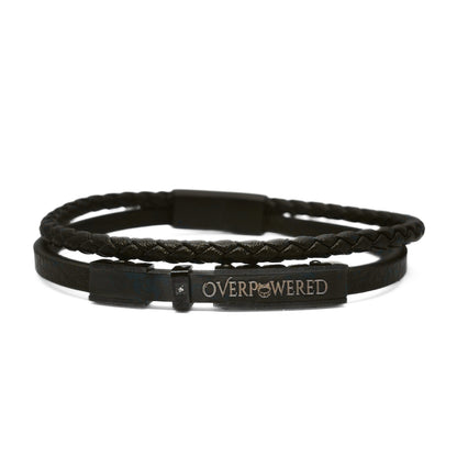 A double-strand black leather bracelet featuring a braided top band and a smooth bottom band with a rectangular metal plaque engraved with the word 'OVERPOWERED' in gold lettering.