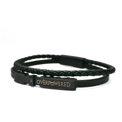 A black double-strand bracelet featuring a braided top layer and a smooth bottom layer with a rectangular metal plate engraved with the word 'OVERPOWERED' in gold.