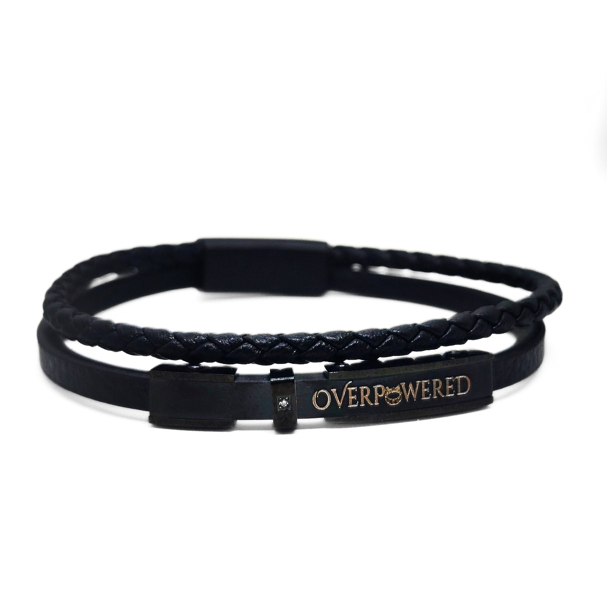 A black double-strand bracelet featuring a braided top band and a smooth bottom band with a rectangular metal clasp engraved with the word 'OVERPOWERED' in gold lettering.