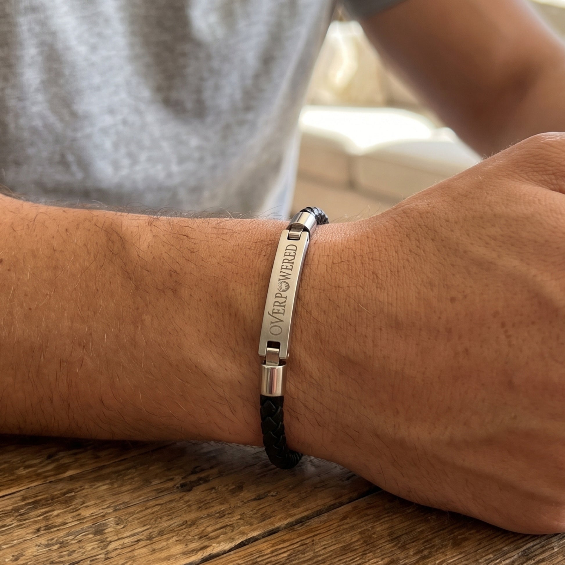 A close-up of a man's wrist wearing a silver-toned metal bracelet engraved with 'OVERPOWERED' and a black braided cord, resting on a wooden table.