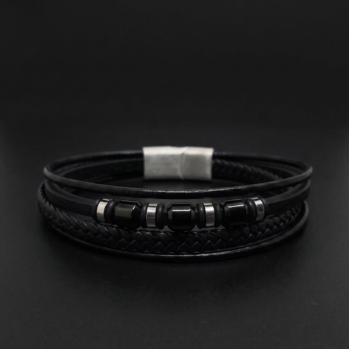A black multi-strand leather bracelet featuring a braided center band, a silver-toned rectangular clasp, and smooth black beads with silver spacer rings, displayed on a dark background.