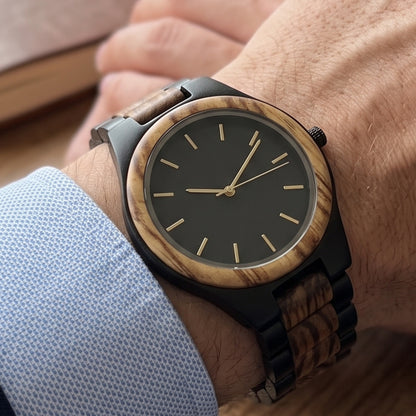 A close-up of a wrist wearing a wooden watch with a black dial and a wood-grain bezel and strap, shown on a person's wrist with a light blue shirt cuff visible.