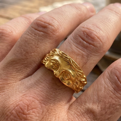 Gold sculpted face ring on hand, wide band with raised eyes and hair relief
