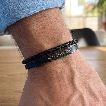 A close-up of a person's wrist wearing a double-strand bracelet. The top strand is a black braided leather, and the bottom strand is a black metal plate engraved with the word 'OVERPOWERED' in gold.