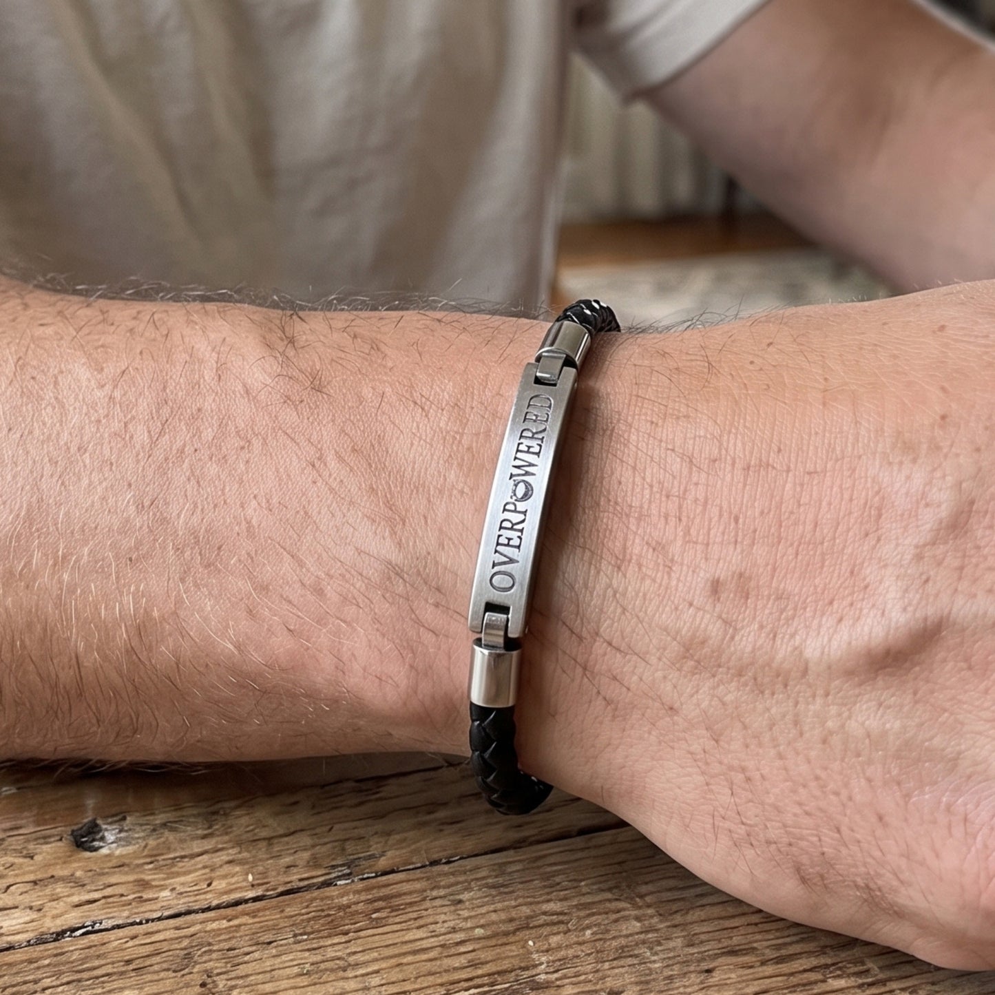A close-up of a person's wrist wearing a black braided cord bracelet with a polished silver metal ID plate engraved with the words 'OVERPOWERED'.