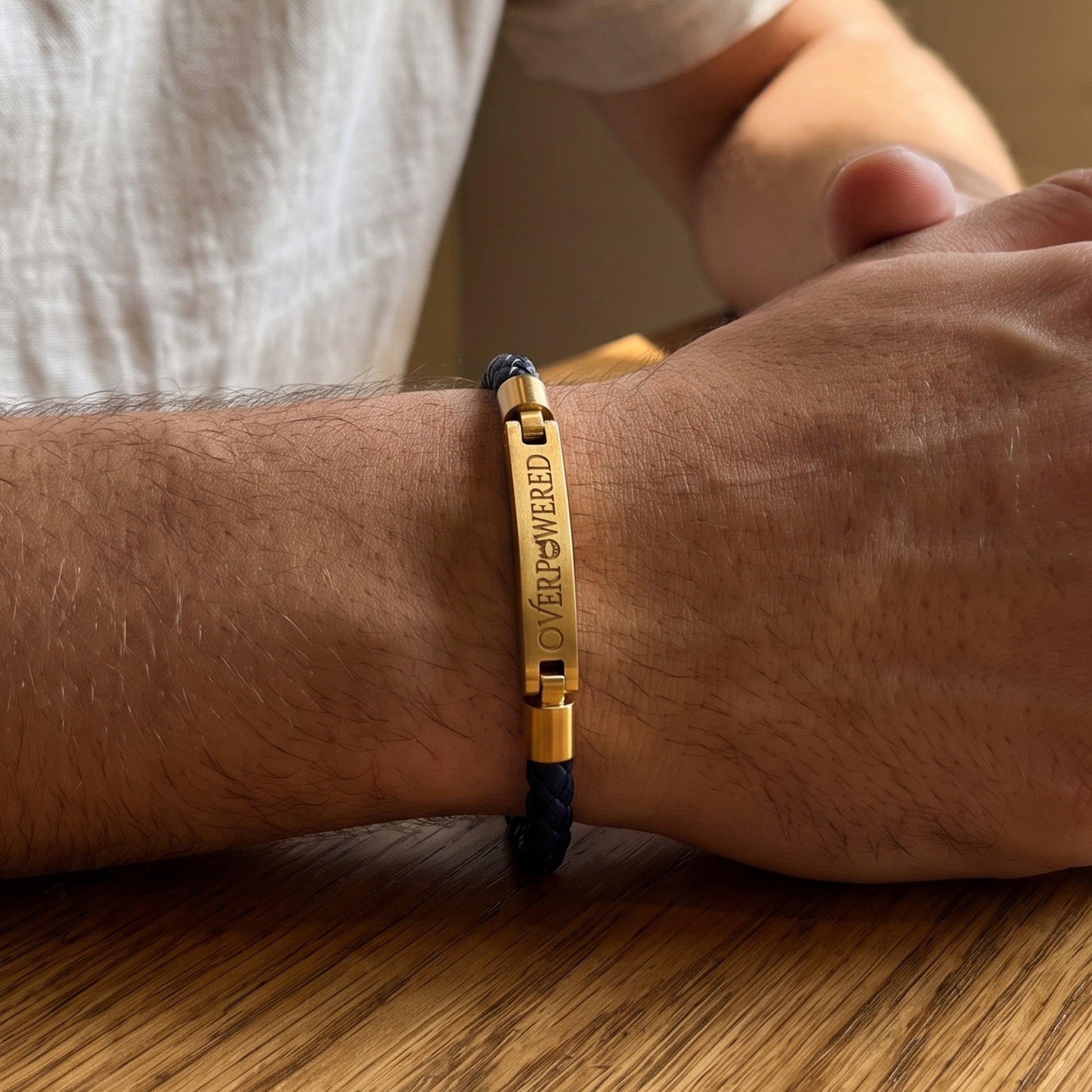 A close-up of a person's wrist wearing a gold-plated bar bracelet engraved with 'OVERPOWERED' on a dark navy braided cord, resting on a wooden table.
