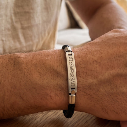 A close-up of a man's wrist wearing a black cord bracelet with a polished silver metal ID plate engraved with the word 'OVERPOWERED'.