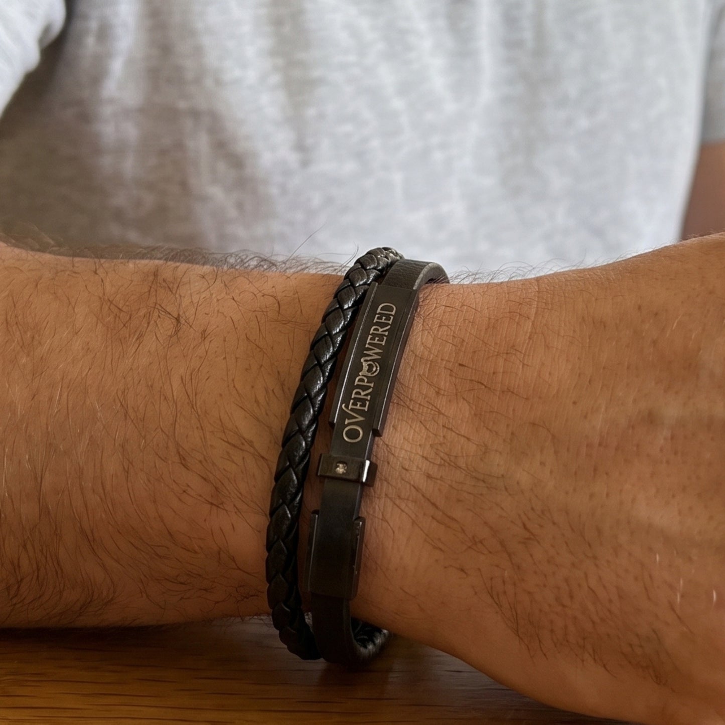 A close-up of a person's wrist wearing two dark bracelets: a braided black leather bracelet and a matte black metal bracelet engraved with the word 'OVERPOWERED'.