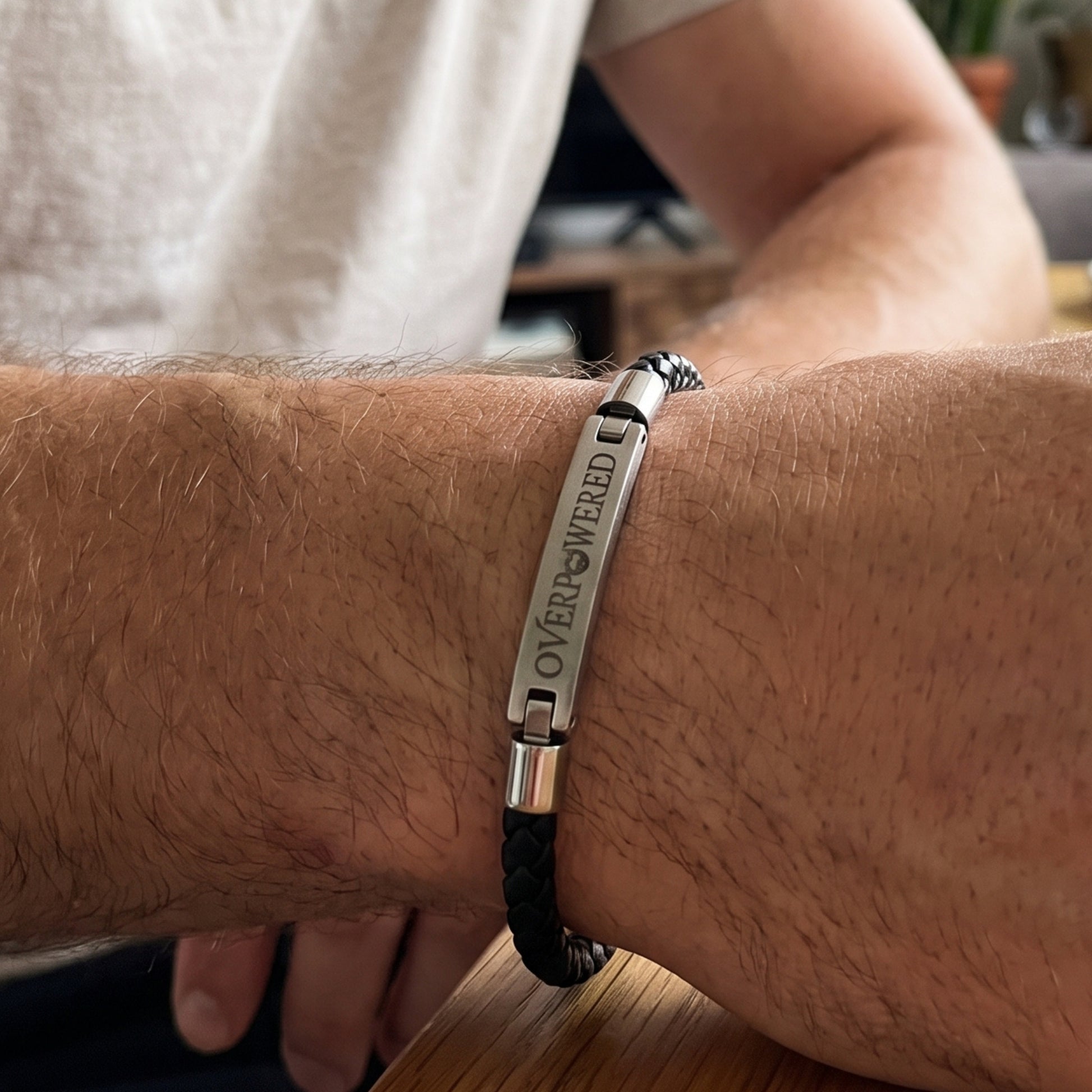 A close-up of a man's wrist wearing a silver-toned stainless steel bracelet with a black braided cord and the engraved text 'OVERPOWERED'.