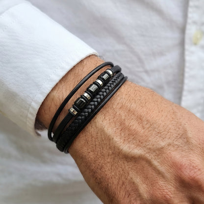 Men's wrist showing black leather wrap bracelet with braided strip and logo beads