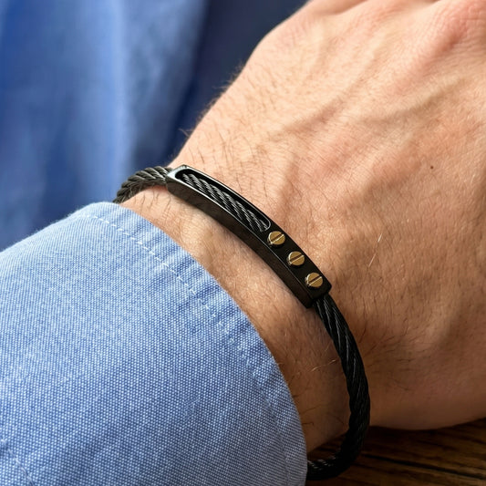 Black braided bracelet with gold accents on a wrist against a blue shirt background
