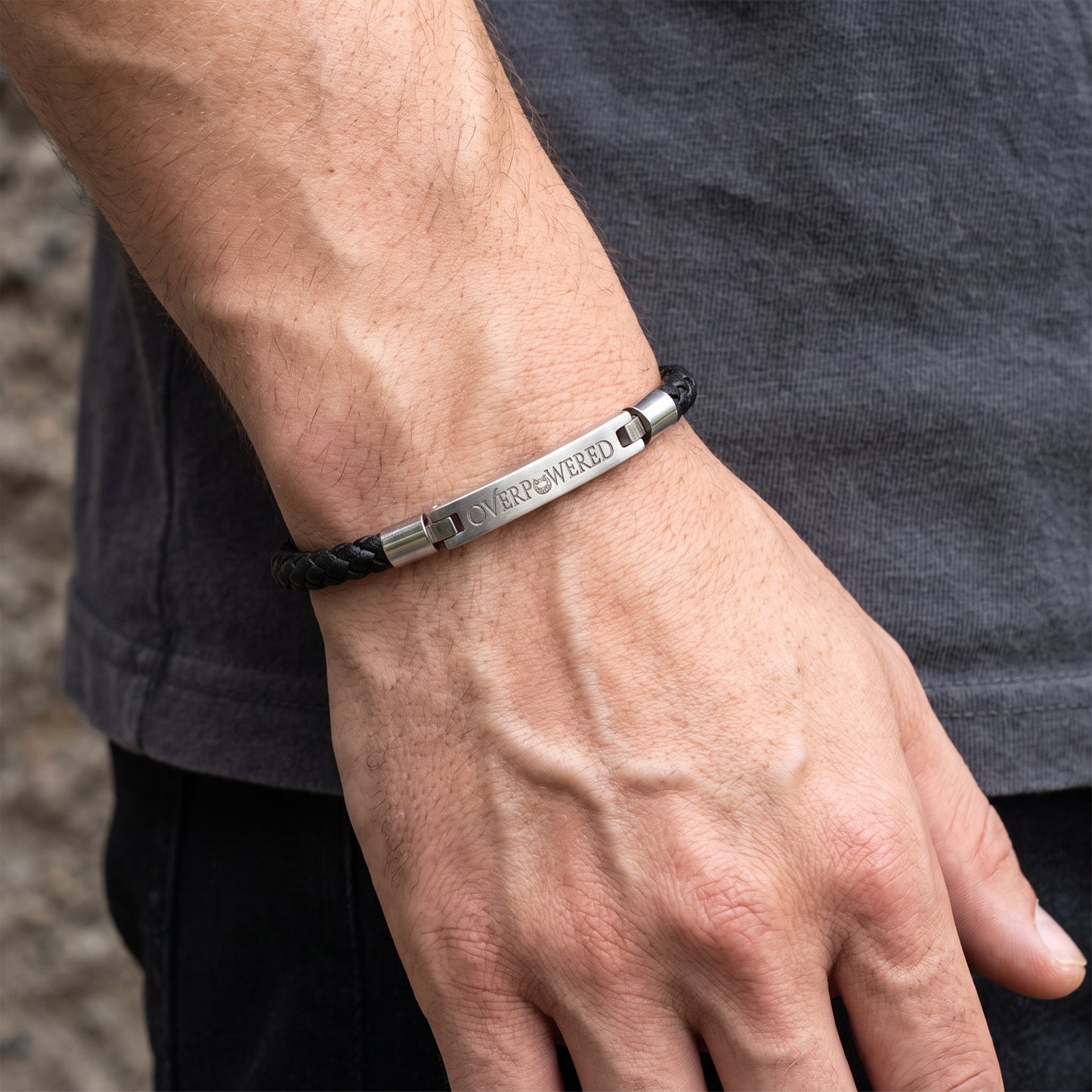 A close-up of a man's hand and wrist wearing a black braided cord bracelet with a polished silver metal bar engraved with the words 'OVERPOWERED'.