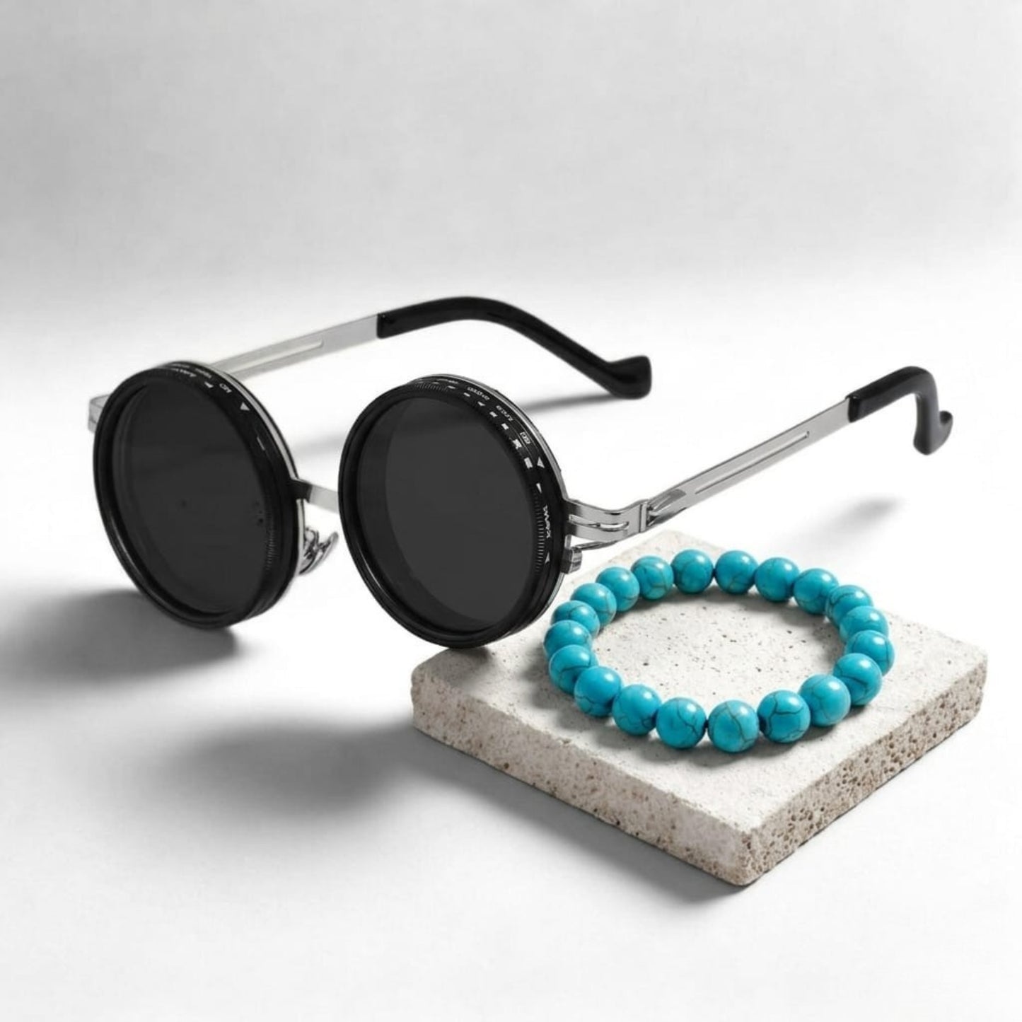Men's sunglasses with round black lenses and silver frames, paired with a turquoise stone bracelet on a light gray surface.