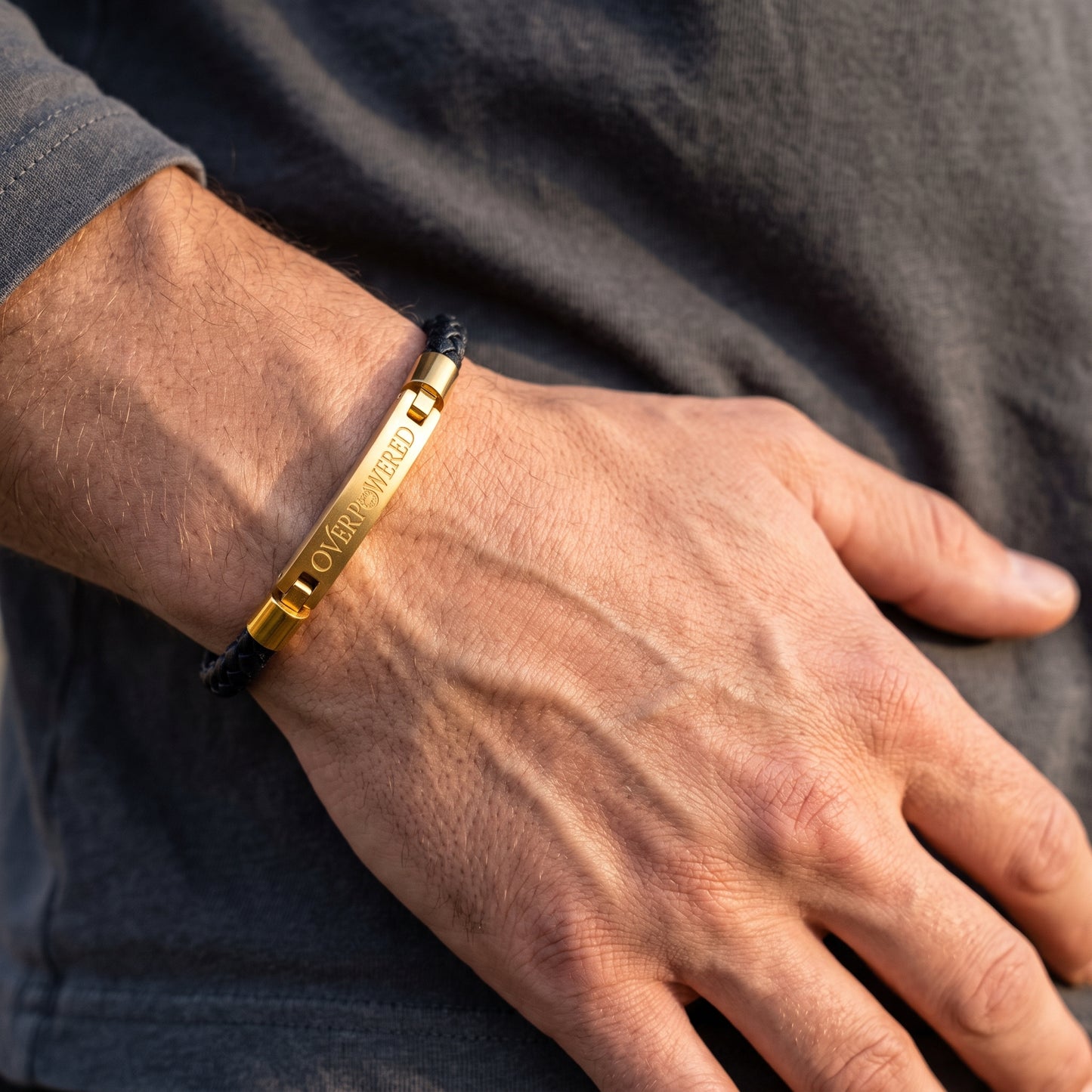 A close-up of a man's hand wearing a gold-plated nameplate bracelet with a black cord, resting on his dark gray shirt. with "OVERPOWERED" on it