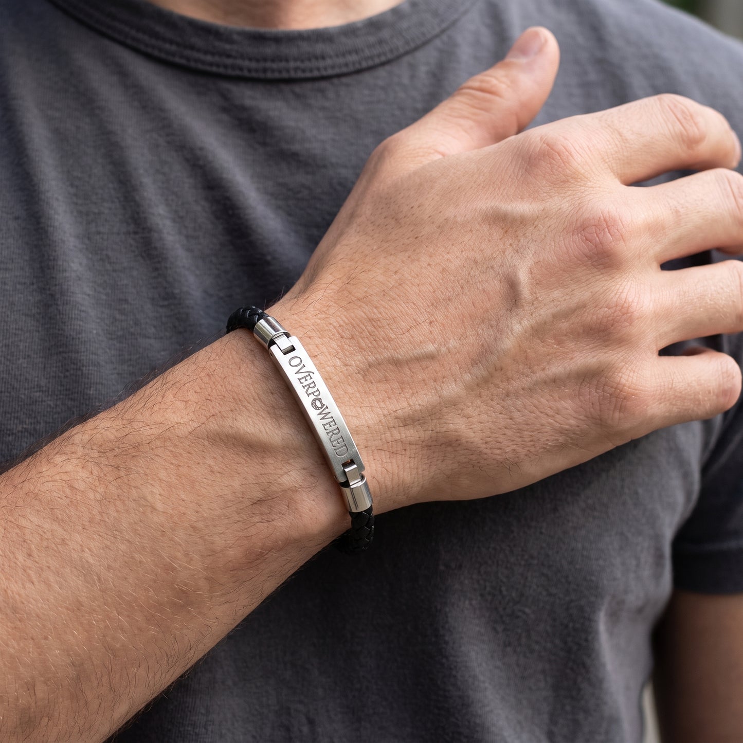 A silver engraved metal bracelet with a black cord, worn on a man's wrist. The bracelet features a polished silver bar engraved with the words 'OVERPOWERED' and 'Eternally Yours' in a vertical orientation, secured with a black cord.