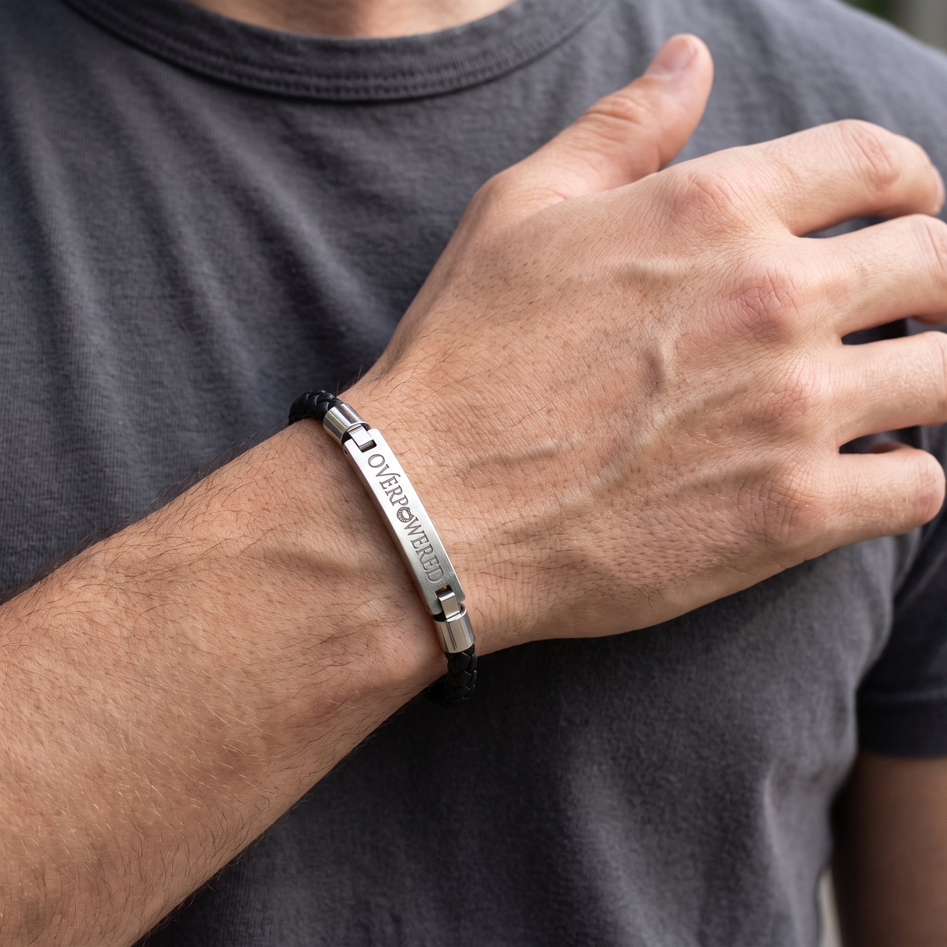 A silver engraved metal bracelet with a black cord, worn on a man's wrist. The bracelet features a polished silver bar engraved with the words 'OVERPOWERED' and 'Eternally Yours' in a vertical orientation, secured with a black cord.