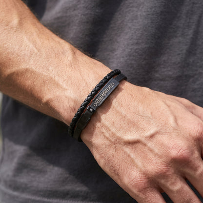 A close-up of a person's wrist and hand wearing two black bracelets, including a braided cord bracelet and a slim metal bracelet engraved with the word 'OVERPOWERED'.