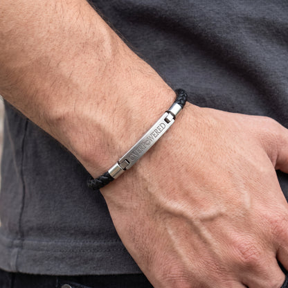 A close-up of a man's wrist wearing a black braided cord bracelet with a polished silver metal plate engraved with the name 'OVERPOWERED'.